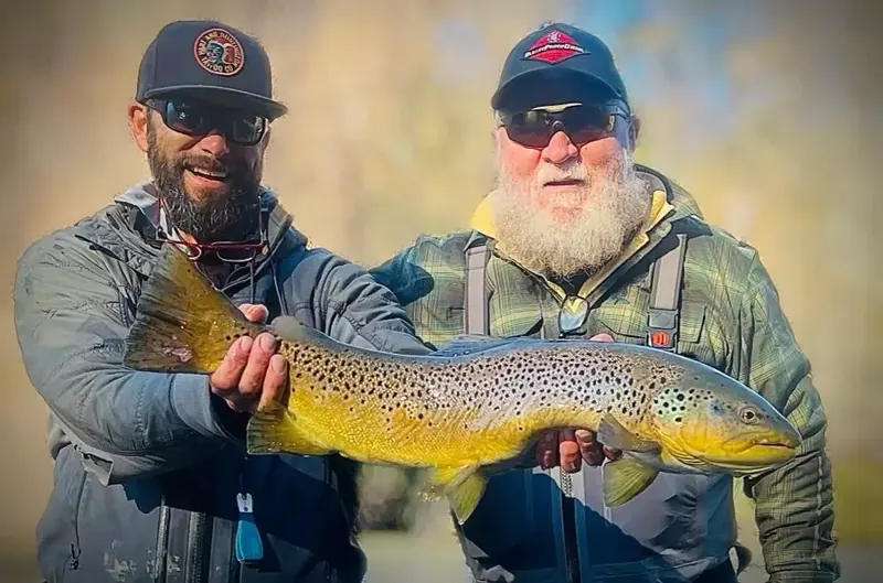 Two smiling anglers holding a large brown trout together