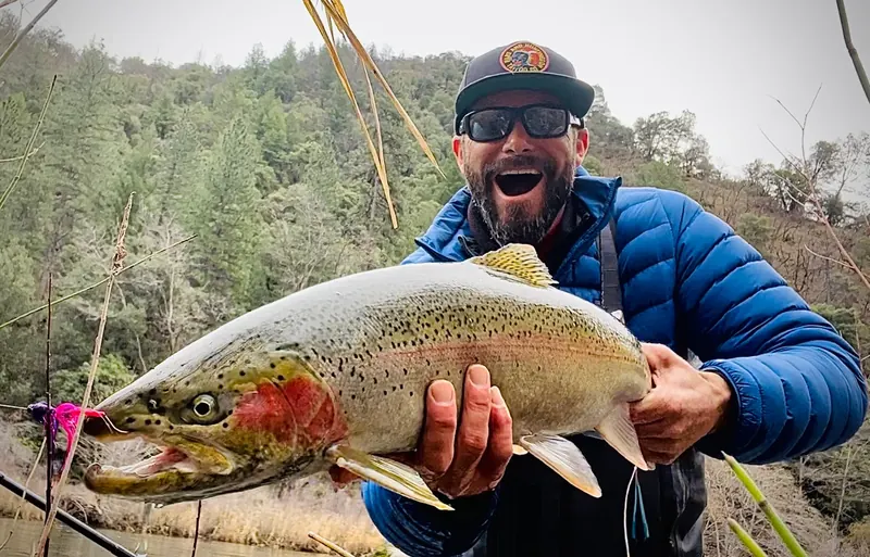 Smiling angler in a blue puffer jacket holding a large chrome steelhead beside a forested Northern California river
